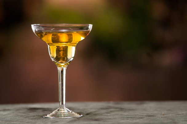 Classy cocktail glass sitting on grey surface, yellow liquid inside with blurry urban background - Zdjęcie, obraz