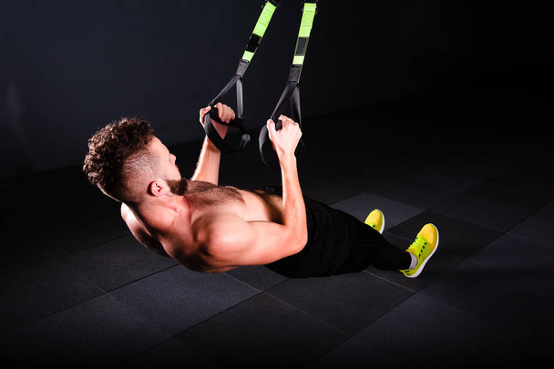One young handsome brave curly bearded muscled man with a beard makes Total Resistance eXercises fitness trx straps working out in gym - Foto, imagen