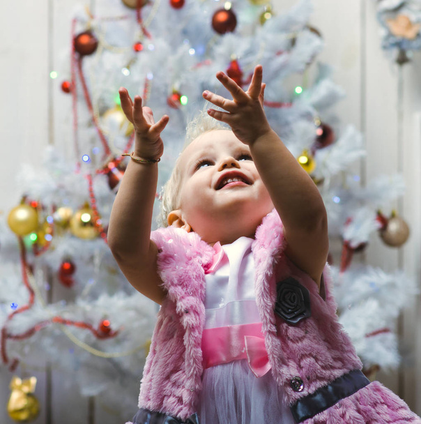 A small child in a beautiful dress stands near a Christmas tree and joyfully catch snowflakes with her hands. - Fotó, kép