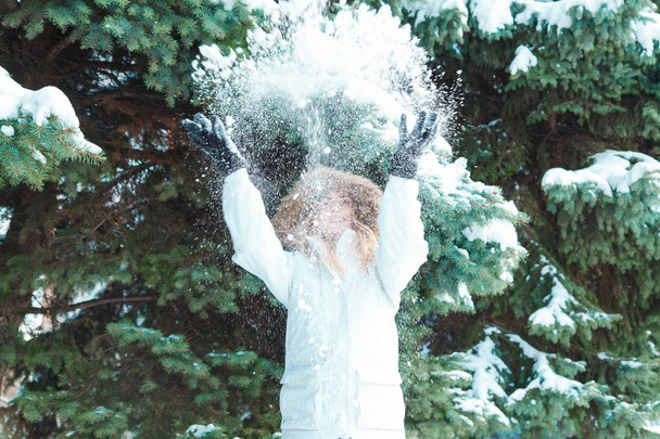 On a cold winter day young beautiful cheerful woman with long blond hair in a white jacket plays with snow against the backdrop of snow-covered trees and cheerfully laughs - Photo, Image