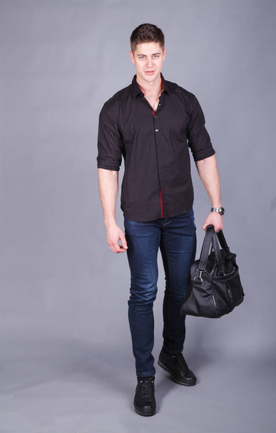 Studio photography, young strong tall, handsome man wearing a black shirt and blue jeans in the hands holding a large black bag - Foto, Bild