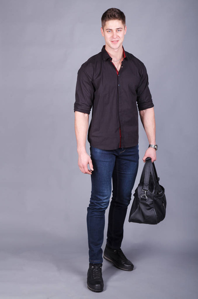 Photographie studio, jeune homme fort grand et beau portant une chemise noire et un jean bleu dans les mains tenant un grand sac noir
 - Photo, image
