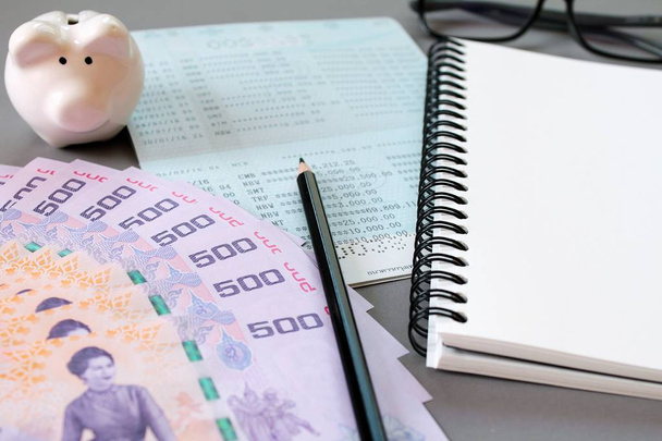 Чистый блокнот, карандаш, сберегательный счет, очки, тайские деньги и копилка на сером фоне
 - Фото, изображение