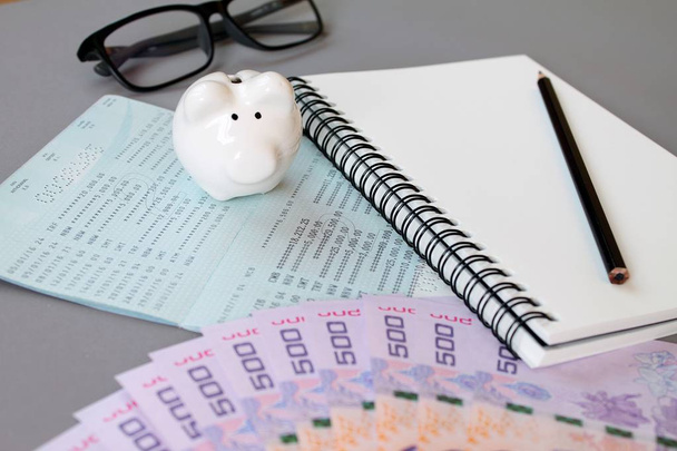 Caderno em branco, lápis, caderno de poupanças, óculos de olho, dinheiro tailandês e banco porquinho em fundo cinza
 - Foto, Imagem