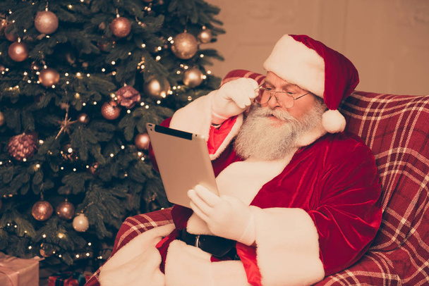 Konzentrierter Weihnachtsmann sitzt im Sessel und liest E-Mails - Foto, Bild