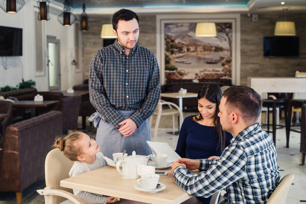 το εστιατόριο και Ενοικιαζόμενα έννοια - σερβιτόρος δίνοντας μενού ευτυχισμένη οικογένεια στην καφετέρια - Φωτογραφία, εικόνα