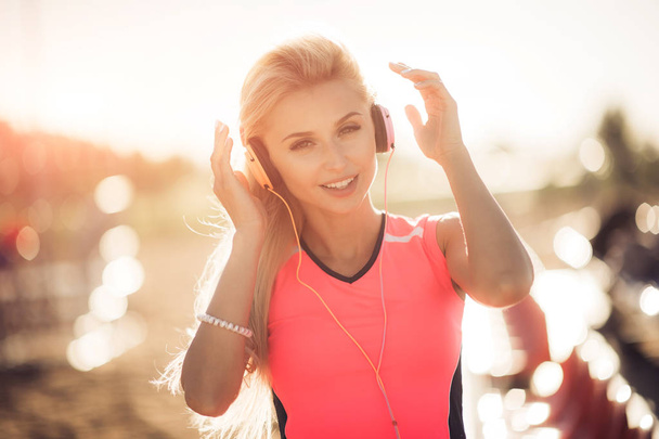 Young woman in sport wear enjoys and smiles listening music with orange earphones. She puts her hands up enjoying life. - Foto, Imagen