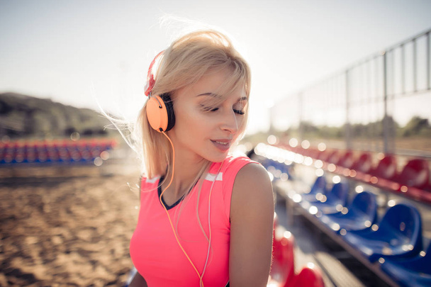 Portrait of a sporty adolescent girl resting from exercising, listening to music with headphones, smiling outdoors. Fitness and sport lifestyle, sunny flare exterior. - Photo, Image