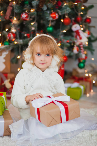 Little beautiful girl sitting near the Christmas tree with gifts in his hands.  in a white sweater and warm headphones - Фото, изображение