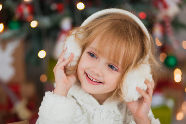 Little beautiful girl in warm white headphones around the Christmas tree - Foto, Imagem