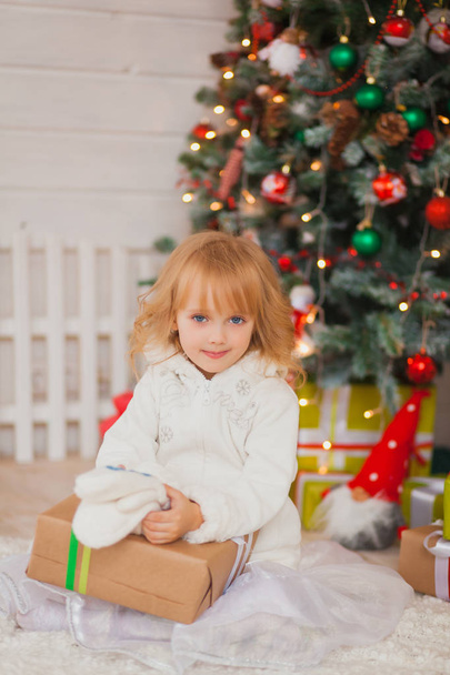 Niña hermosa sentada cerca del árbol de Navidad con regalos en sus manos. en un suéter blanco y auriculares cálidos
 - Foto, imagen