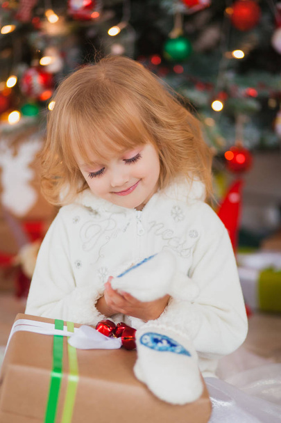 Little beautiful girl sitting near the Christmas tree with gifts in his hands. in a white sweater and warm headphones - Zdjęcie, obraz