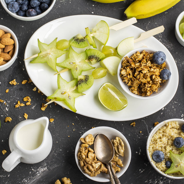 Healthy fresh breakfast. Granola, quinoa bowl, various nuts and fruit skewers with green fruits on a dark background. Top view. Color year. Greenery - Фото, изображение