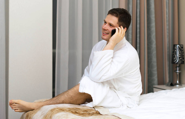 glücklicher Mann im Bademantel mit Handy im Hotelzimmer. - Foto, Bild