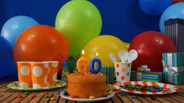 Birthday cake on rustic wooden table with background of colorful balloons, gifts, plastic cups and plastic plate with candies and blue wall in the background - Photo, Image
