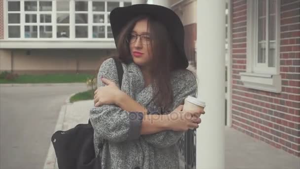 Ładna kobieta w czarny kapelusz i okulary z kubkiem kawy idąc ulicą - Materiał filmowy, wideo