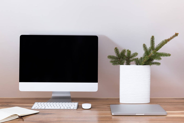 Modern workspace. Computer, laptop and a notebook on a wooden walnut table with new year composition A vase with a fir branch for christmas mood. - 写真・画像