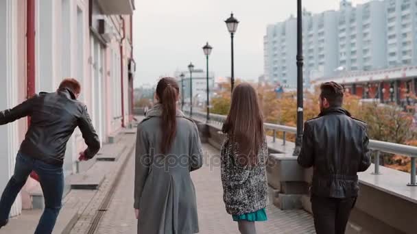 Two trendy girls and two stylish men cheerfully walk in the city, jump and have fun. Slow mo, steadicam shot, back view - Video