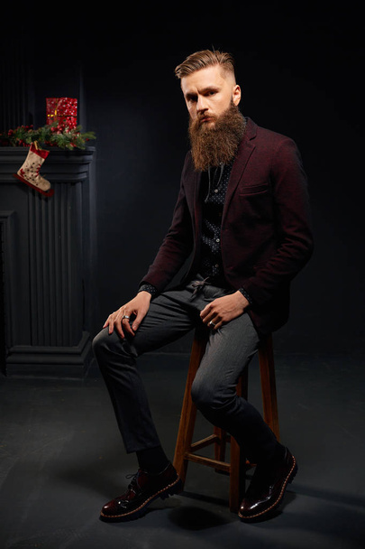 Handsome stylish man with beard sitting in dark studio with christmas sock on background - Valokuva, kuva