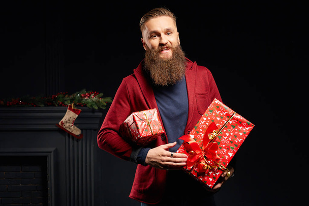 Young smiling man with long beard with red christmas boxes in his hands - Photo, Image