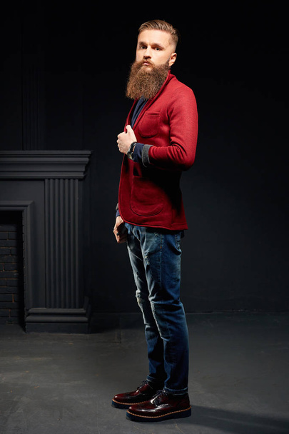 Attractive model male with long beard in dark studio wearing red jacket - Фото, изображение