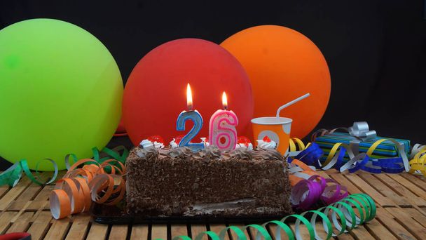 Pastel de cumpleaños de chocolate con velas ardiendo en la mesa de madera rústica con fondo de globos de colores, regalos, tazas de plástico y serpentinas con fondo negro
 - Foto, imagen