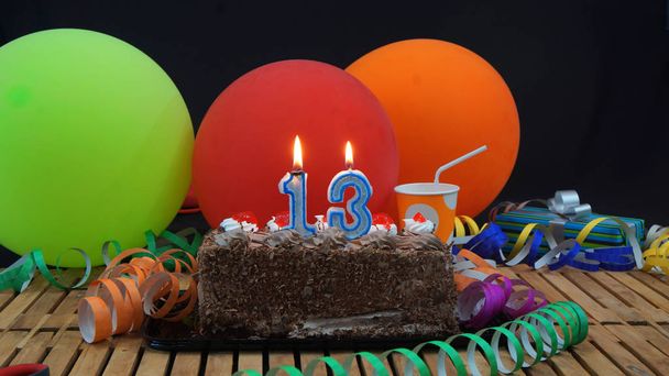 Pastel de cumpleaños de chocolate con velas ardiendo en la mesa de madera rústica con fondo de globos de colores, regalos, tazas de plástico y serpentinas con fondo negro
 - Foto, imagen