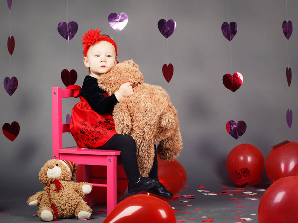 Portrait of white Caucasian cute adorable little baby girl toddler sitting on small pink chair with bear toy in studio wearing red dress and headband, holiday Valentine day - Foto, Imagem
