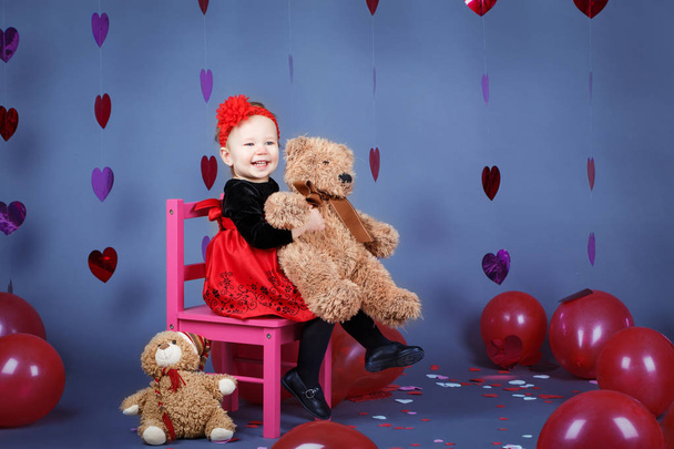 休日バレンタイン、鉢巻き笑って楽しんで笑顔赤いドレスを着てスタジオでクマのおもちゃと小さなピンクの椅子に座って白い白人かわいい愛らしい赤ちゃん女の子幼児の肖像画 - 写真・画像