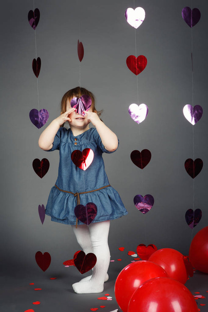 Portrait of cute adorable funny white Caucasian little girl toddler in studio with red balloons hearts on grey background making faces having fun, Valentine holiday, happy childhood concept - Fotografie, Obrázek