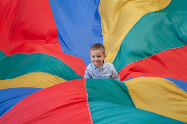 Portrait of white Caucasian child boy toddler sitting in the center of playground rainbow parachute celebrating his birthday at party - Photo, Image
