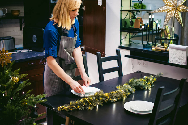 жінка домогосподарка встановлює стіл. молода блондинка в сукні і фартусі, подається в обідньому столі, кладе посуд, робить роботу на кухні
 - Фото, зображення