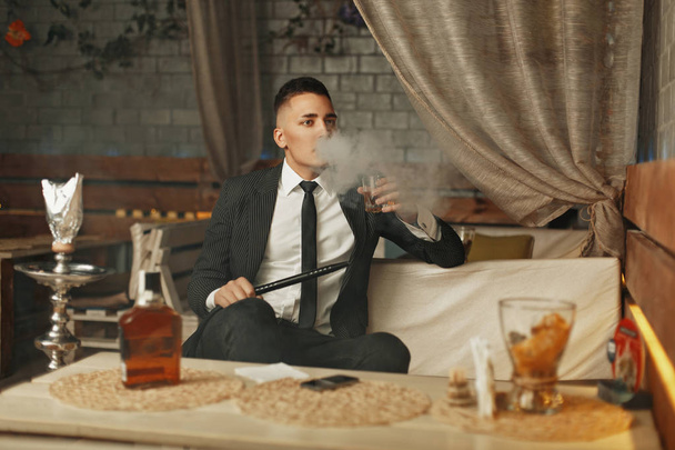 Young rich man in an elegant suit has a rest in a bar and smokes a hookah. - Фото, зображення