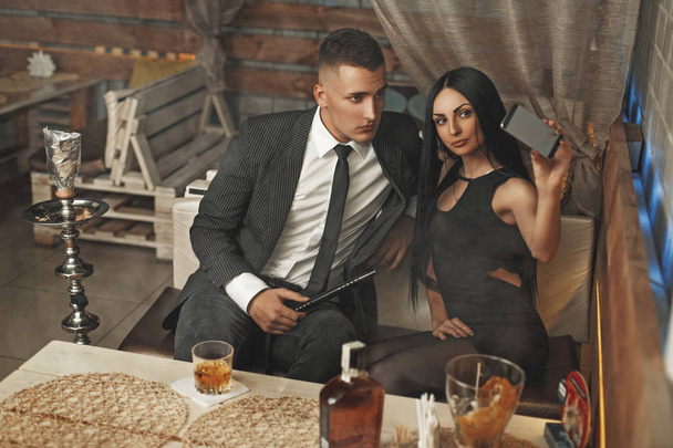 Beau jeune couple élégant se détendre dans un bar et fume un narguilé. Sont photographiés eux-mêmes au téléphone
 - Photo, image