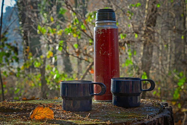 garrafa térmica desgastado vermelho com duas xícaras azuis em um tronco de árvore
 - Foto, Imagem