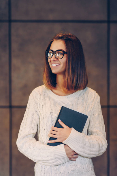 Beautiful smiling girl in stylish glasses and a white sweater with sketchbook on the background of a concrete wall - Фото, изображение
