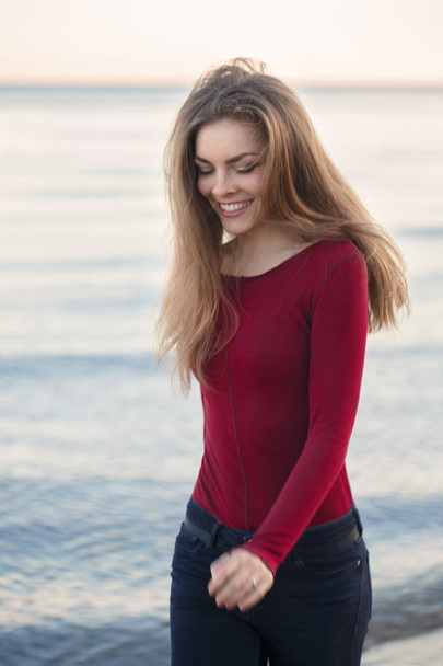 Retrato de estilo de vida de la joven mujer delgada caucásica con el pelo largo desordenado usando jeans negros y camisa roja caminando en el día ventoso al aire libre en la orilla de la playa al atardecer, desenfoque de acción de movimiento
 - Foto, imagen