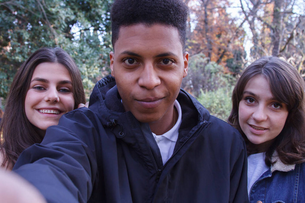 Група молодих людей, дві кавказькі дівчата і чорний хлопчик роблять селфі в полі з мобільним телефоном
. - Фото, зображення