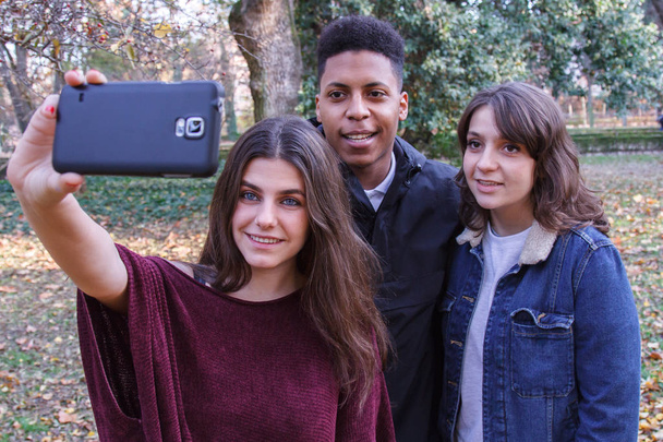 Ryhmä nuoria, kaksi valkoihoista tyttöä ja musta poika tekemässä selfietä kentällä kännykällä.
. - Valokuva, kuva