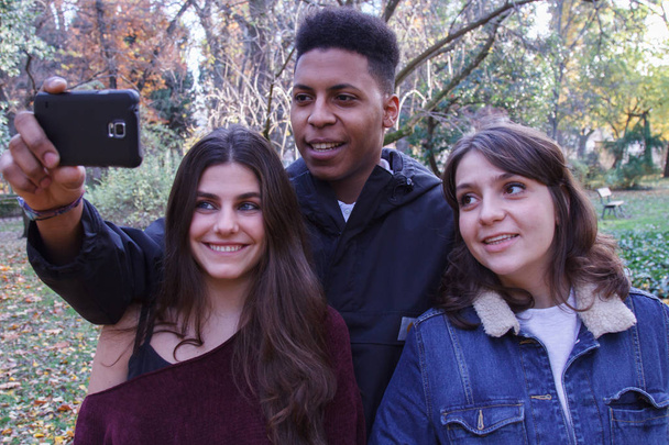 Grupo de jóvenes, dos chicas caucásicas y un chico negro haciendo una selfie en el campo con un teléfono celular
. - Foto, Imagen