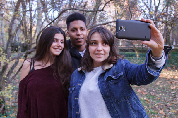 Csoport fiatal, két kaukázusi lány és egy fekete fiú, csinál egy selfie, egy sejt telefon területén. - Fotó, kép