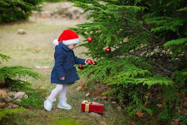 little girl in Santa hat decorates a Christmas tree in the forest - Foto, imagen