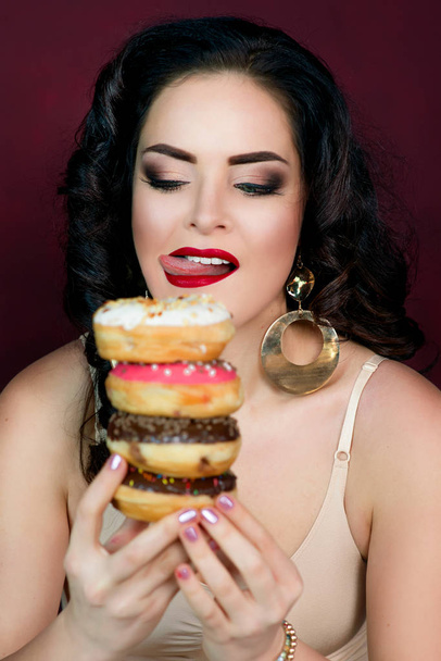 young, beautiful, attractive, darkhaired woman with colorful, tasty, glazed doughnuts on marsala background - Foto, afbeelding