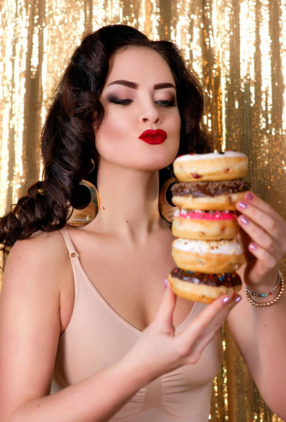 young, beautiful, attractive, darkh aired woman with colorful, tasty, glazed doughnuts on gold background - Fotó, kép
