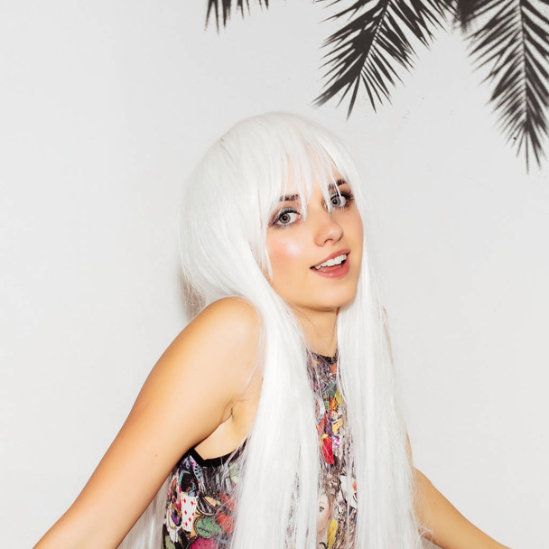 Beautiful summer woman pointing to her teeth with palm tree background. Cheerful funny playful young cute girl in colorful clothes and white wig show how to smile with perfect teeth. Fun hipster girl. - Photo, image