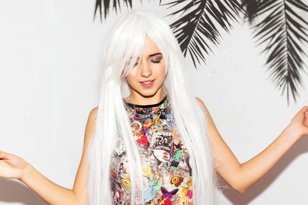 Beautiful summer woman with palm tree background. Cheerful funny playful young cute girl in colorful clothes and white wig. Fun hipster girl. Calm meditation balance and equilibrium concept. - 写真・画像
