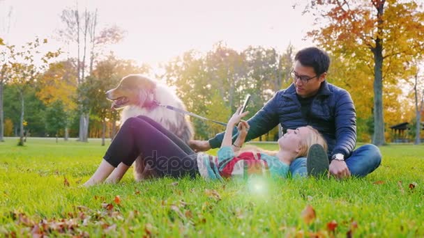 Mezirasový sex mladý pár odpočívá v parku se psem. Asiat hladil psa, běloška, čtení e knihy čtenář nebo tablet používá - Záběry, video
