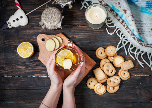 female hands holding cup black tea with lemon, ginger and cookies on dark wood - Fotó, kép