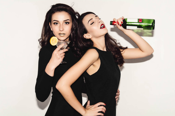 Two beautiful women in black night fashion dress posing isolated on a white gray background. Pretty brunette girl friends twins having fun drinking cocktails. Singing and dancing. Bottle of wine. - Foto, Imagem
