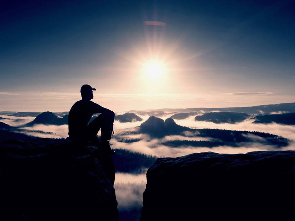 Sylwetka turysta sam czarno-cap siedzieć na klifie. Niebieski filtr Fotografia. Człowiek chce mis - Zdjęcie, obraz